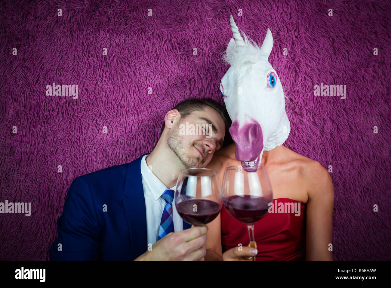 Portrait of unusual couple on purple background. Freaky young woman in comical mask drinking wine with guy in suit. Young man with girl unicorn. Stock Photo