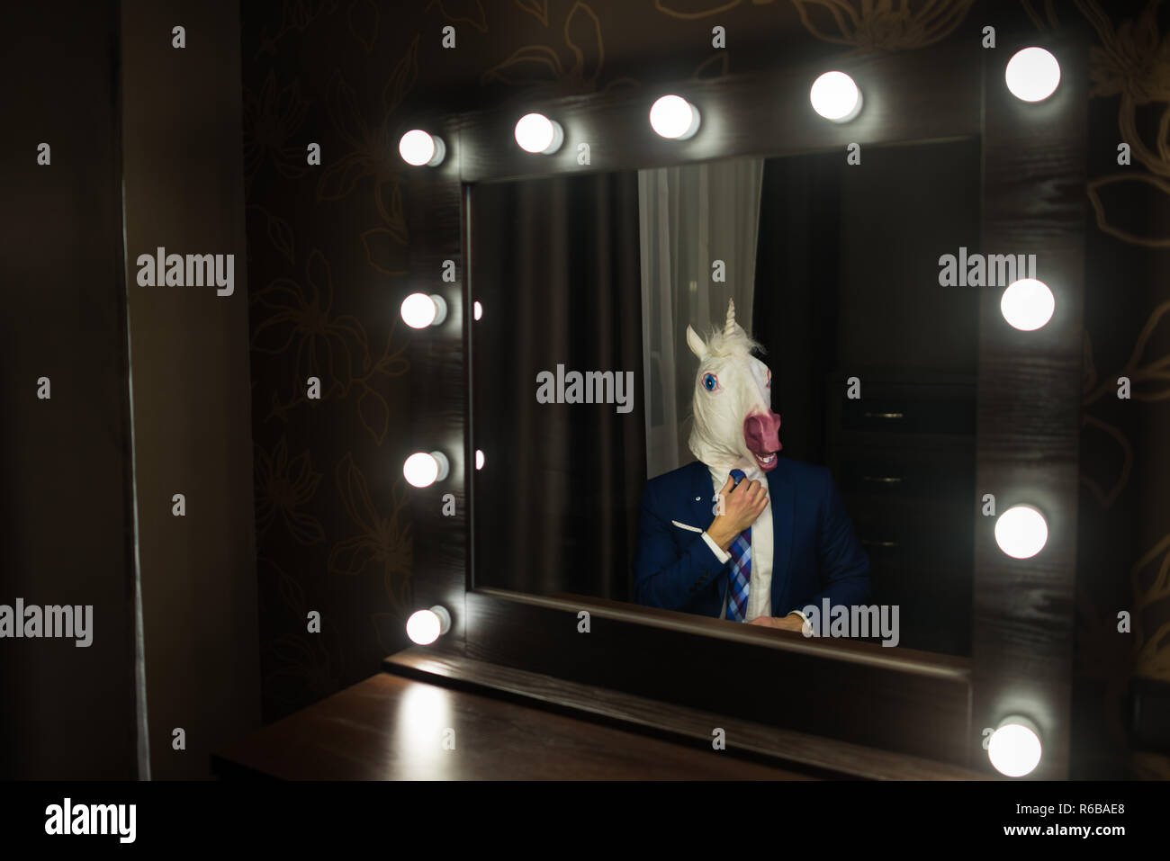 Glamour man in elegant suit and funny mask corrects tie and looks at himself in mirror in dressing room. Freaky guy posing like boss. Unusual unicorn. Stock Photo
