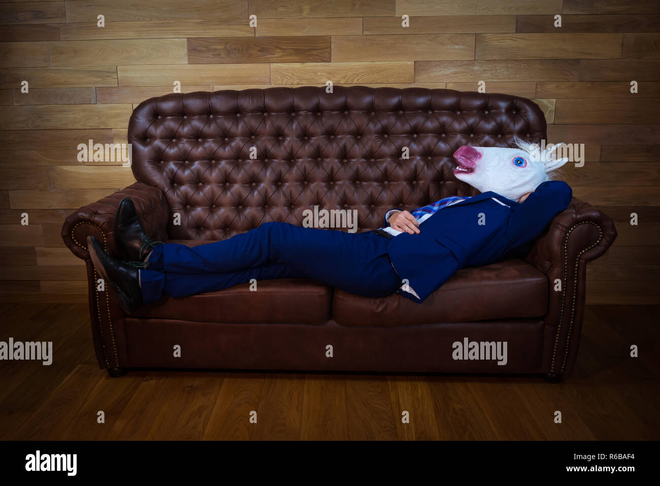 Funny unicorn in elegant suit lies on leather sofa. Unusual man relaxing at home. Freaky guy in comical mask on background of wooden wall. Stock Photo