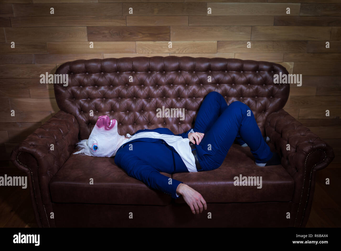 Funny unicorn in elegant suit lies on sofa. Unusual man at home. Freaky guy in comical mask on background of wooden wall. Stock Photo