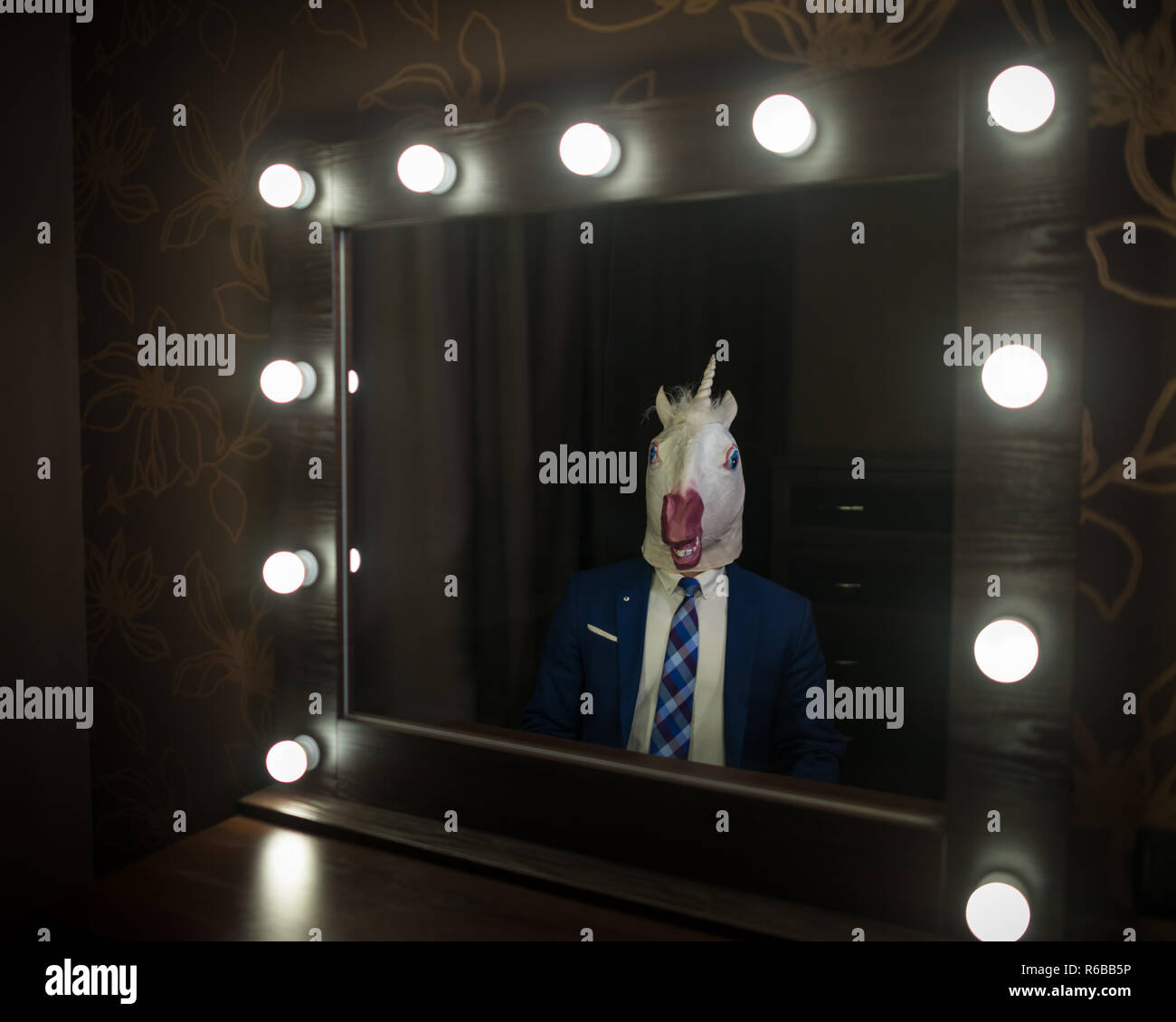 Glamour guy in suit and mask looks at himself in the mirror in dressing room. Freaky young man in stylish room posing like a boss. Unusual unicorn. Stock Photo