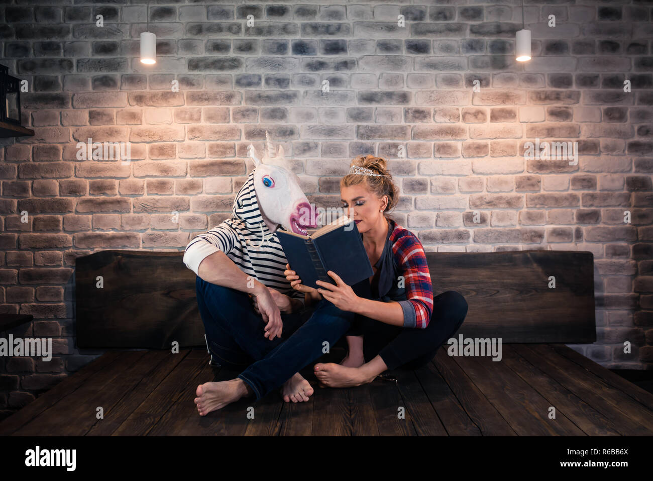 Unusual couple reading a book in stylish room. Beautiful girl sits on bed with funny boyfriend in comical mask. Young woman with freaky man. Stock Photo