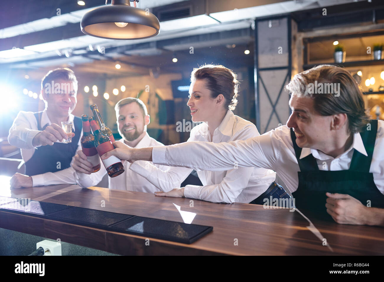 Happy excited young restaurant colleagues reaching hands with beers to toast for good job, they sitting at at bar counter Stock Photo