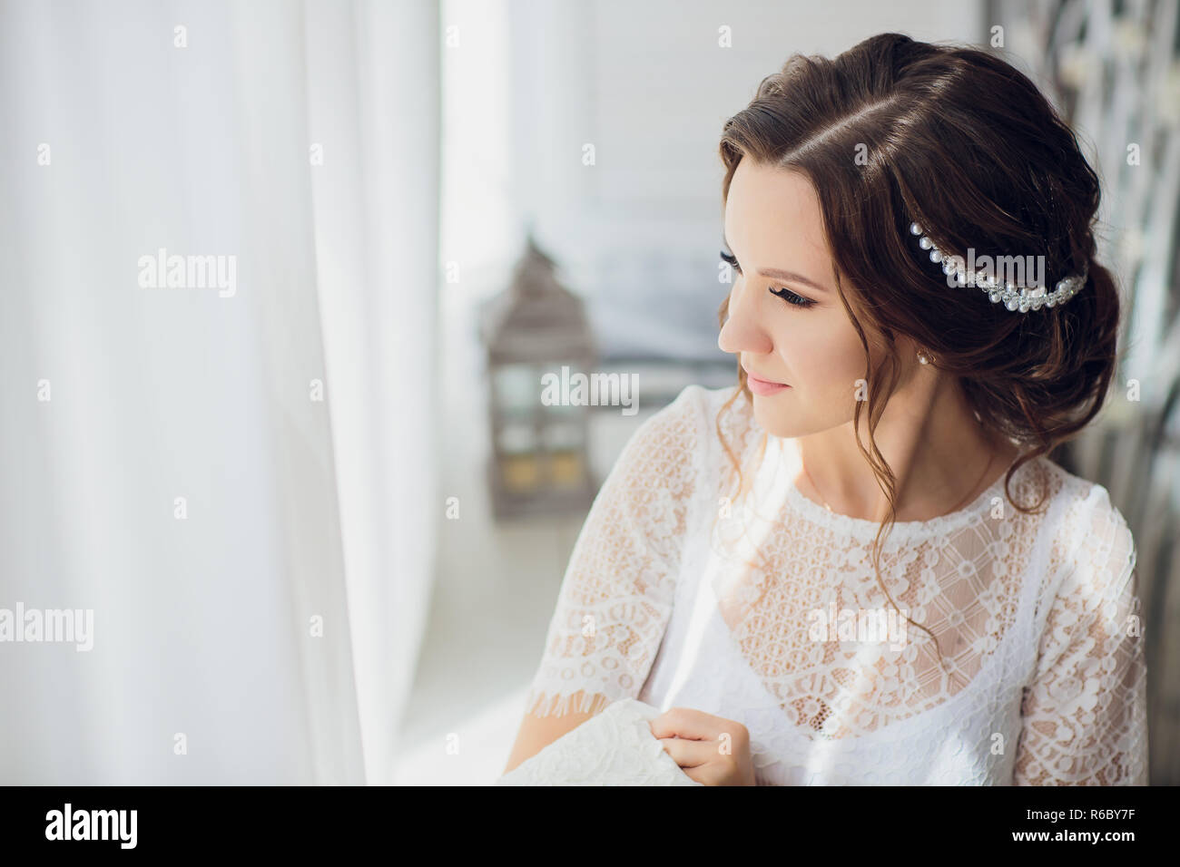 Pretty brunette bride in silk dressing gown and lacy veil holding wedding  dress on a hanger near the window Stock Photo - Alamy