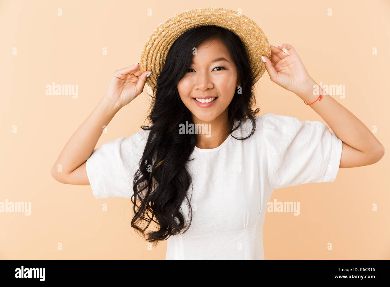 Portrait of a happy young asian woman in straw hat isolated over beige ...