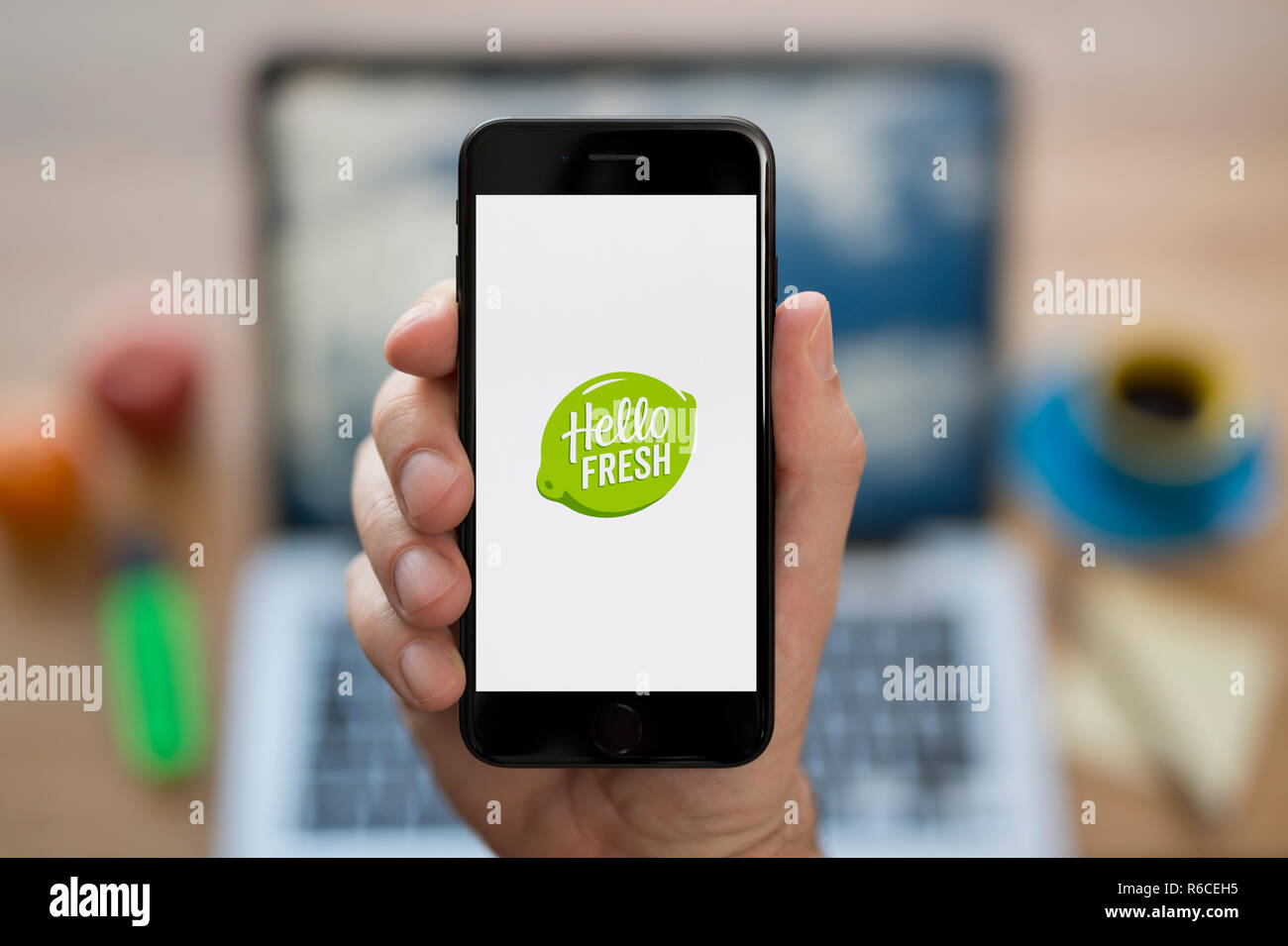 A man looks at his iPhone which displays the Hello Fresh logo, while sat at his computer desk (Editorial use only). Stock Photo