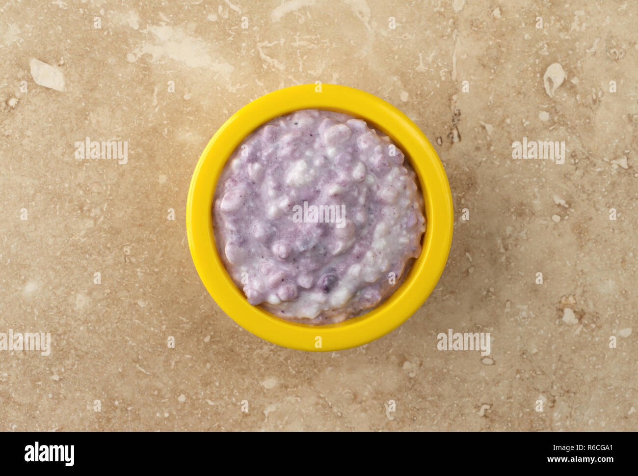 Top view of a small bright yellow bowl filled with blueberries and cottage cheese on a beige mottled tile. Stock Photo
