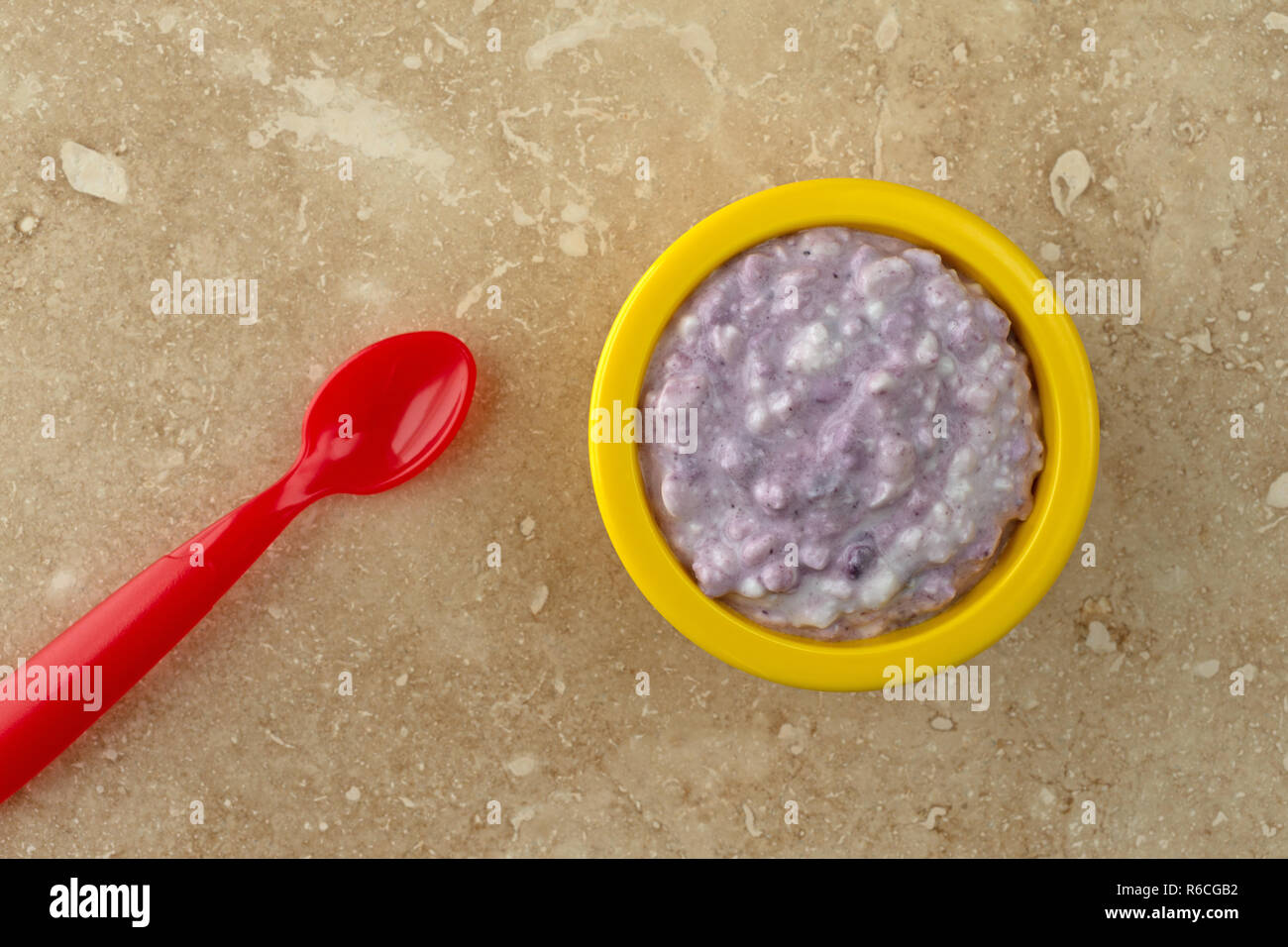 Overhead view of a small yellow bowl filled with blueberries and cottage cheese plus a red spoon atop a beige mottled tile. Stock Photo