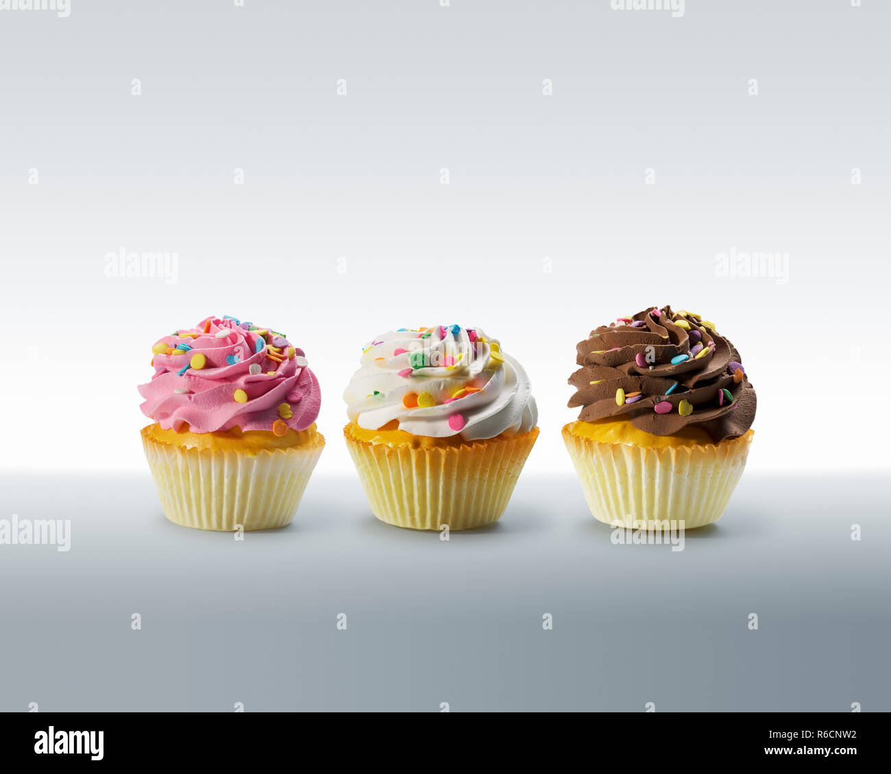 Three cupcakes with frosting and colourful sweets on a plain surface Stock Photo