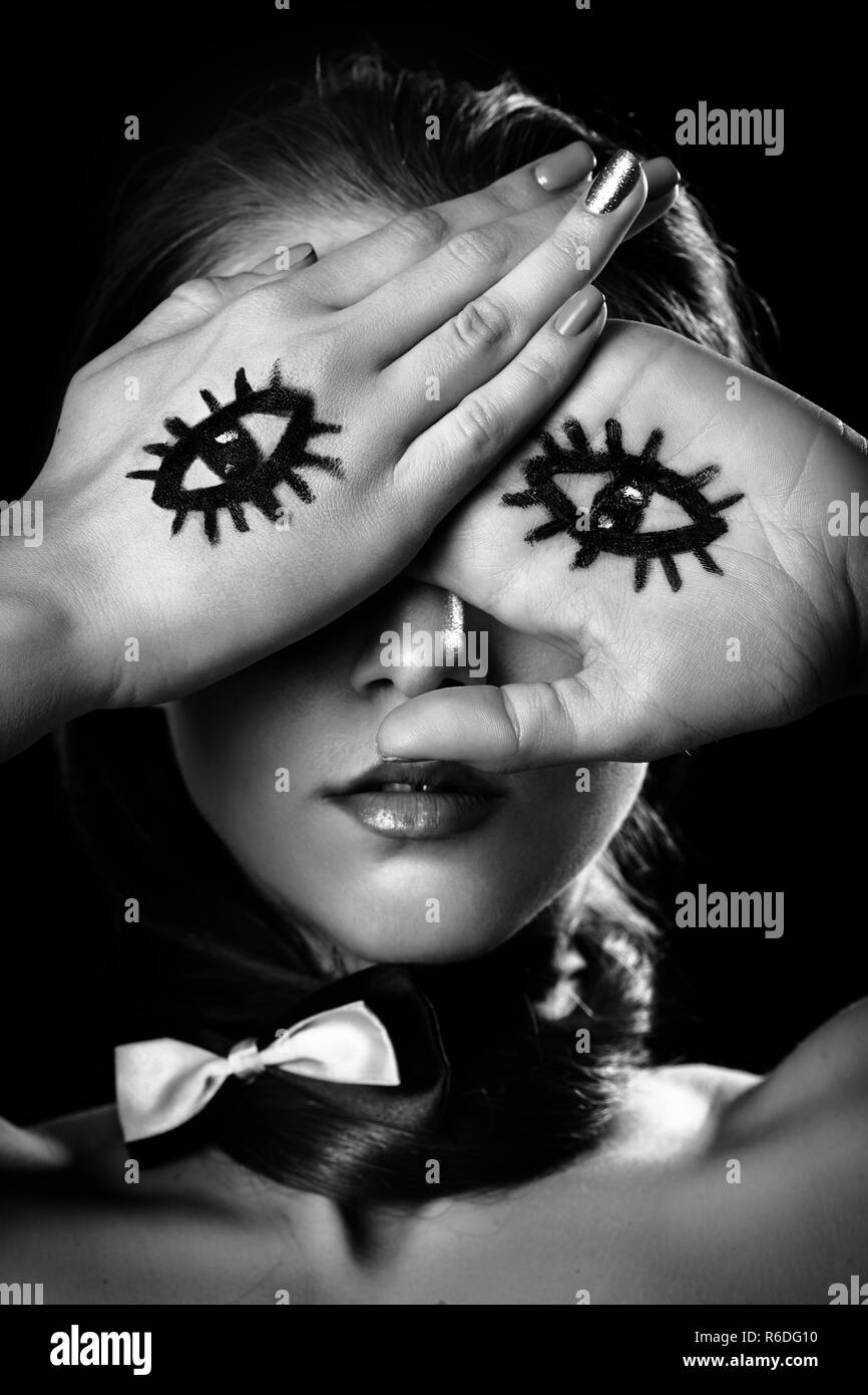 young woman with tie butterfly cover face with palms with drawn eyes, black background, monochrome Stock Photo