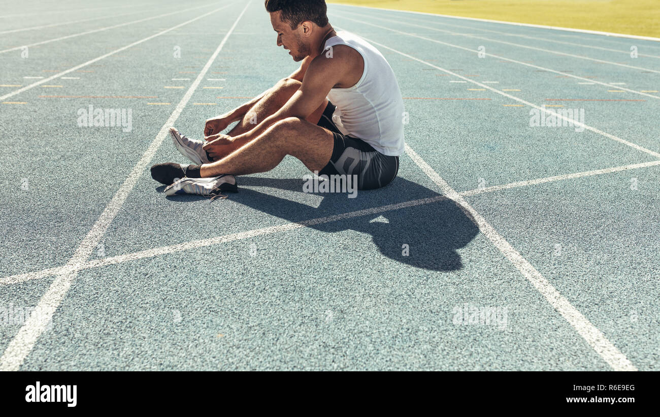 Athlete sitting on a running track tying shoe lace. Runner sitting on the running track wearing his shoes. Stock Photo