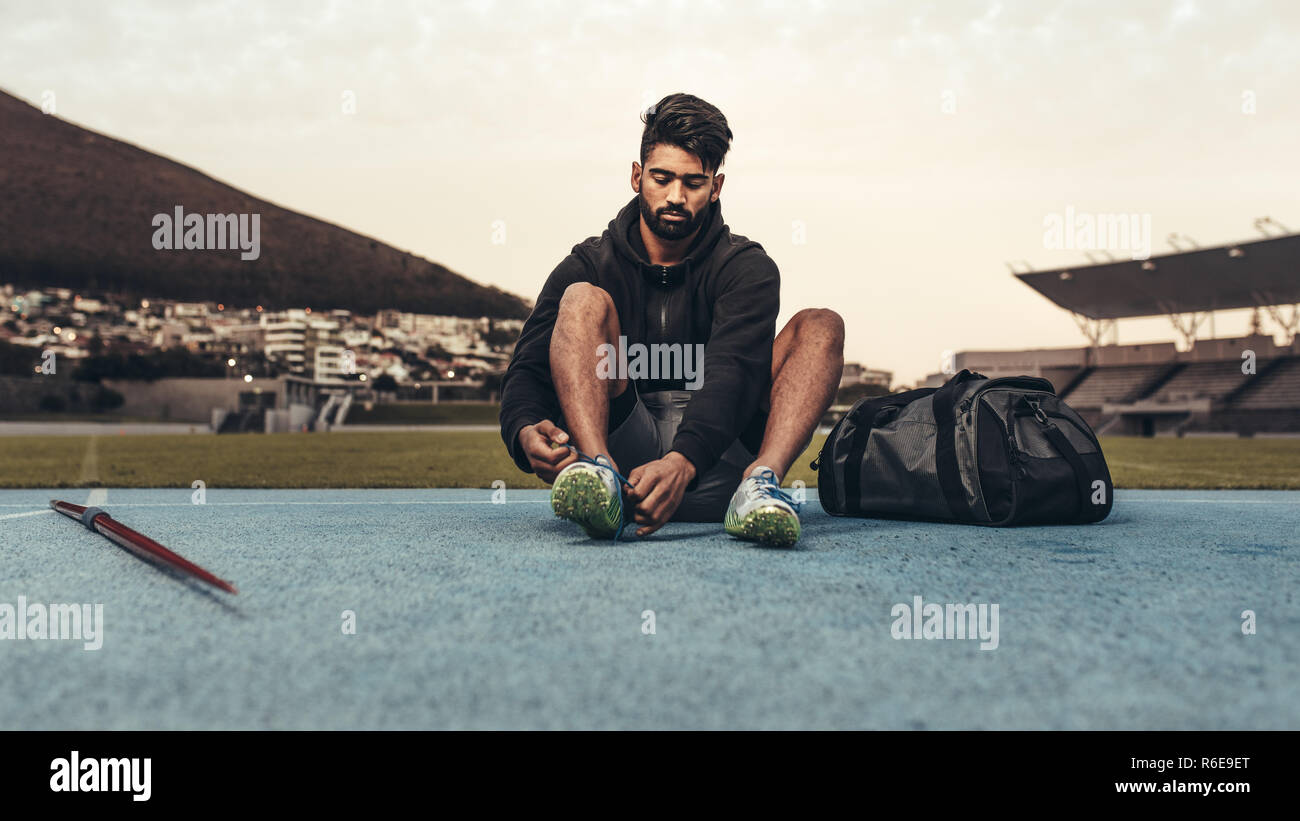 Athlete getting ready for training wearing shoes sitting in a track and field stadium. Man tying shoe lace sitting on track with a javelin and bag by Stock Photo