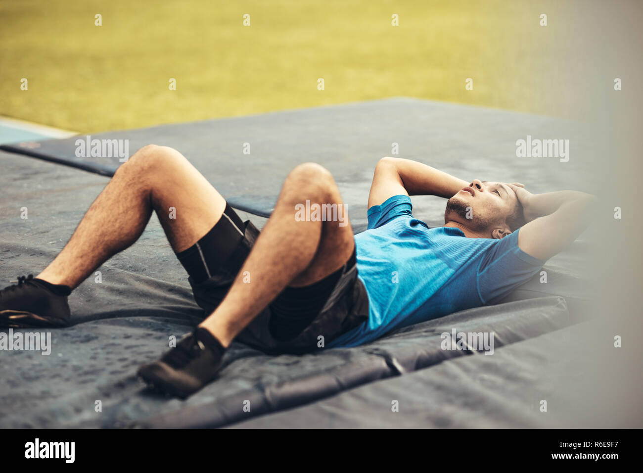 Athlete lying on high jump landing mat looking tired. Athlete holding his hands to his head lying on landing mat after a high jump. Stock Photo