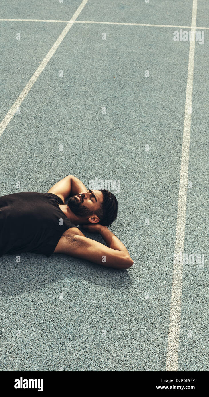 Runner lying on the track in a relaxed mood with hands under his head. Top view of an athlete relaxing after a run. Stock Photo