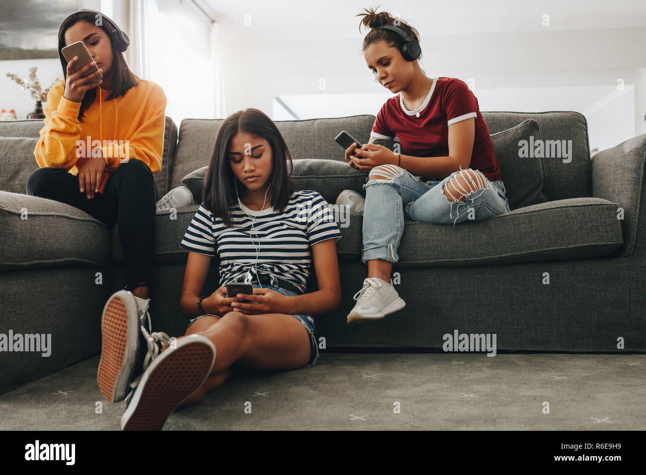Three girls sitting at home listening to music independently using headphones. Friends sitting at home and checking their mobile phones. Stock Photo