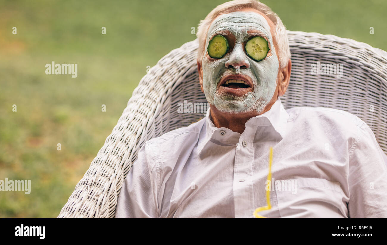 Retired man sitting on chair with clay facial mask and cucumber slice on face. Senior man taking facial spa treatment. Stock Photo