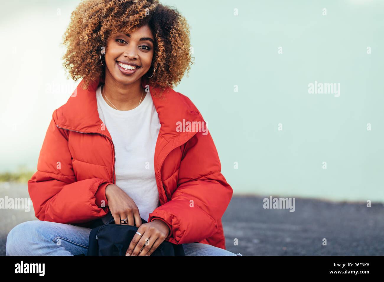 Cheerful afro american woman sitting outdoors on street. Smiling tourist woman in jacket sitting on street holding a bag. Stock Photo