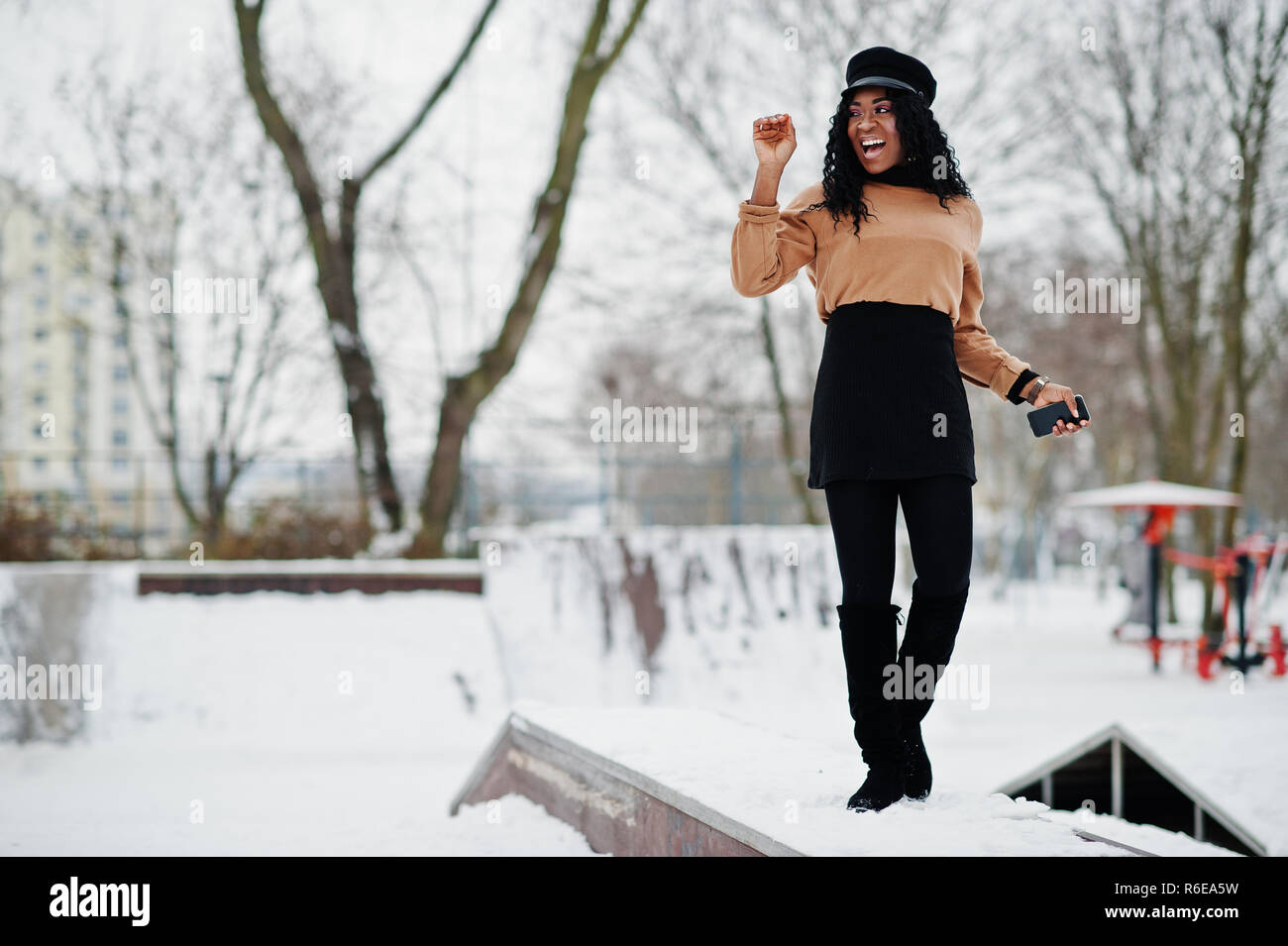 African american woman in black skirt, brown sweater and cap posed at  winter day against snowy background, holding phone at hand Stock Photo -  Alamy