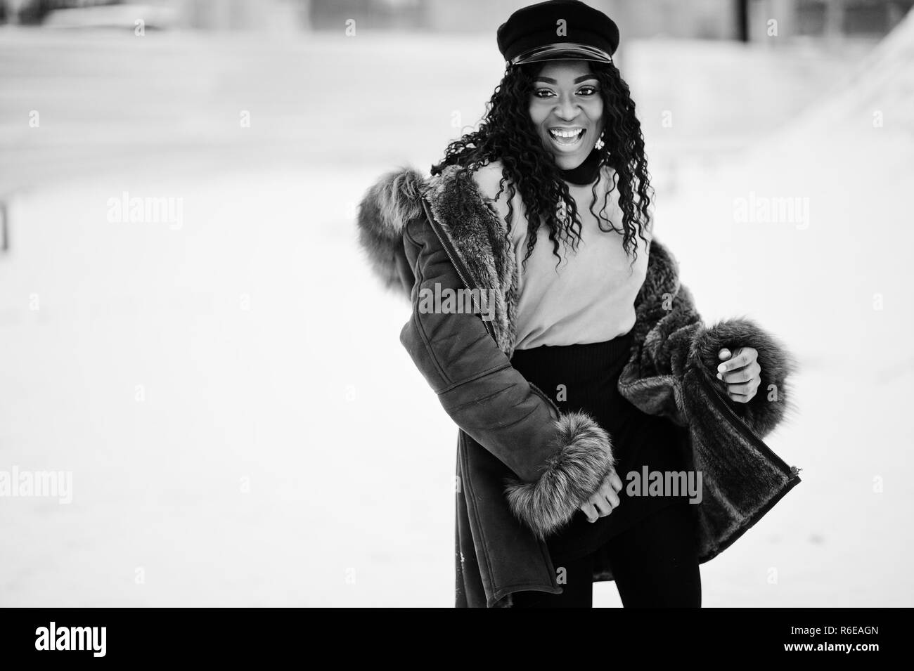 African american woman in sheepskin coat and cap posed at winter day against snowy background. Stock Photo