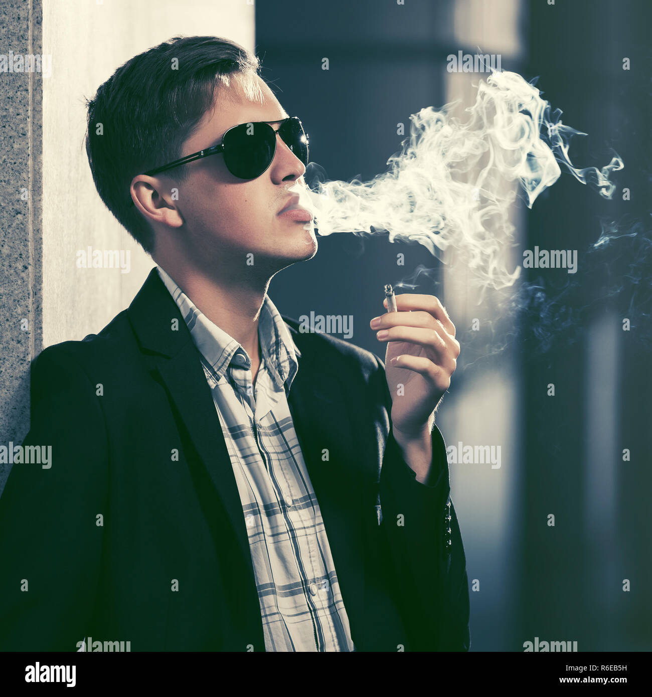 Young handsome man in sunglasses smoking a cigarette in city street. Stylish fashion male model in black blazer Stock Photo