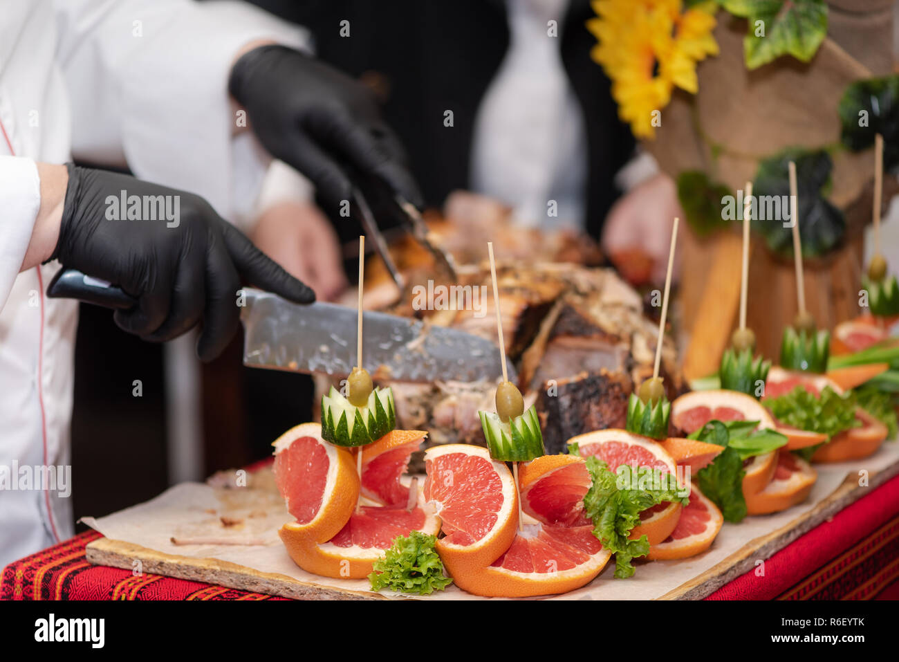 Chief cook cuts into pieces freshly cooked pork hip, process close up. Decorations of fruits and vegetables on the table, catering. Show at restaurant Stock Photo