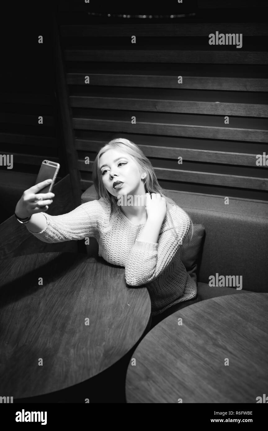 young blond woman in a knitted sweater, making a selfie photo on a smartphone, sitting at a table in a cafe, spending time alone, doing internet communication Stock Photo