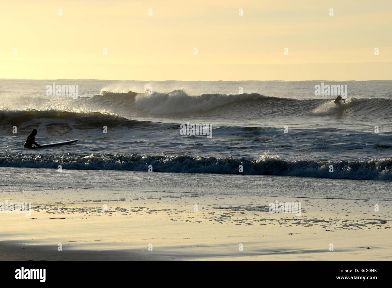 A surfer gets to his feet on an early morning wave with a friend about to join him in the shallows Stock Photo