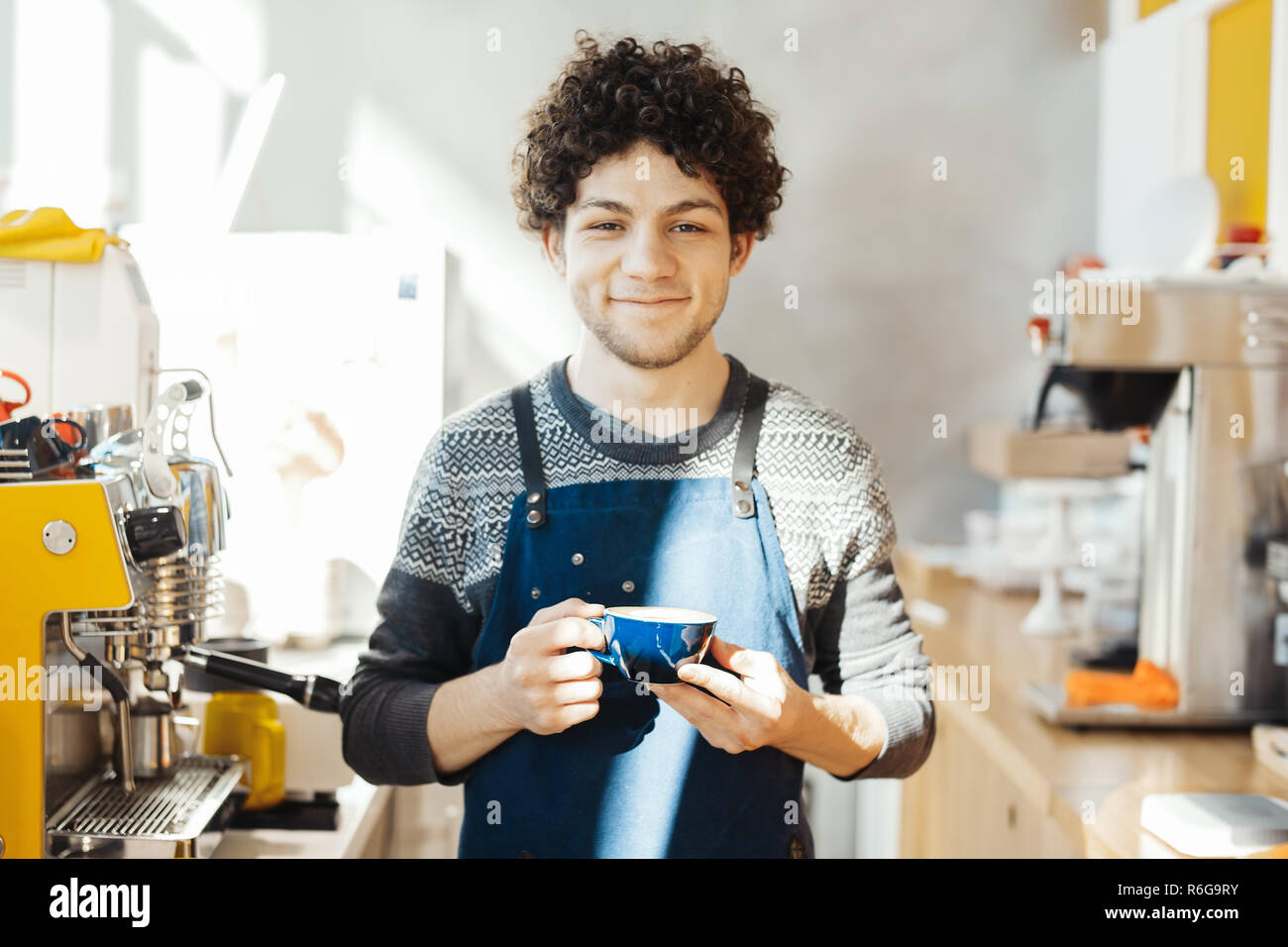Barista smiling and holding coffee cup near bar counter in bright