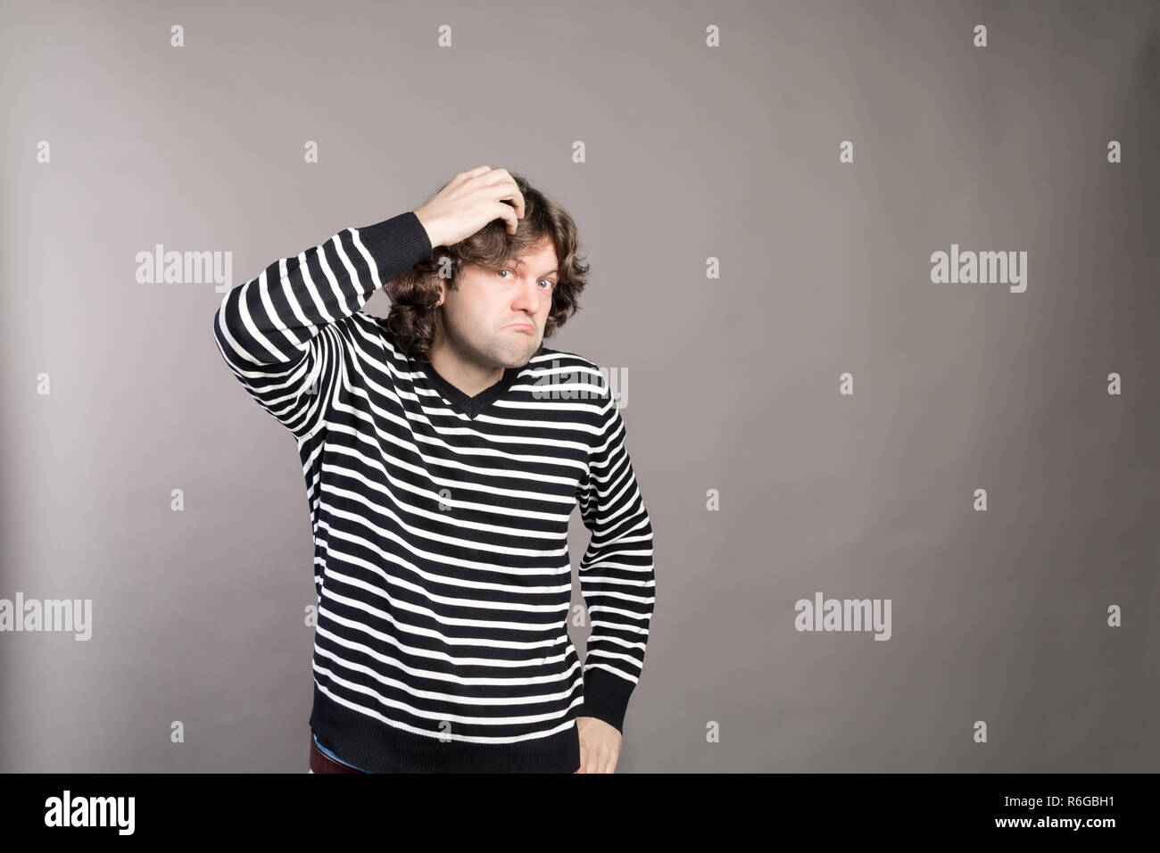 Puzzled guy with messy hair, frowning and looking unsatisfied scratching head, thinking deeply about something on grey wall background. Human facial expression, emotion, feeling, sign body language Stock Photo