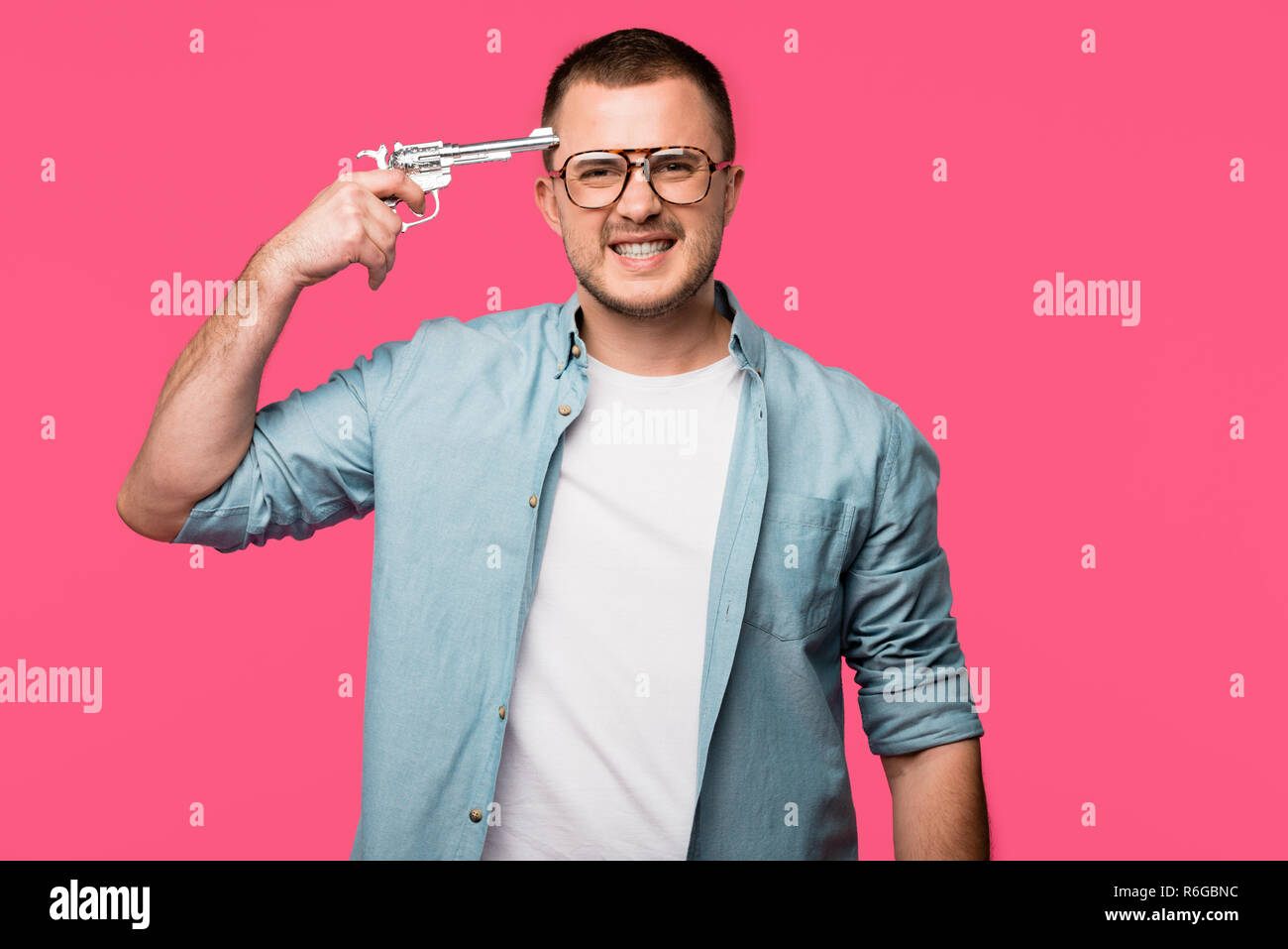 young man in eyeglasses holding revolver near head and looking at camera isolated on pink Stock Photo