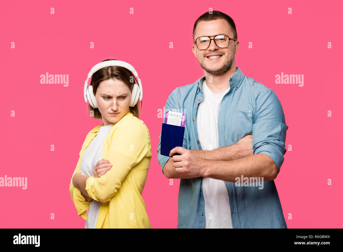 upset woman in headphones looking at happy man in eyeglasses holding passport and boarding pass isolated on pink Stock Photo