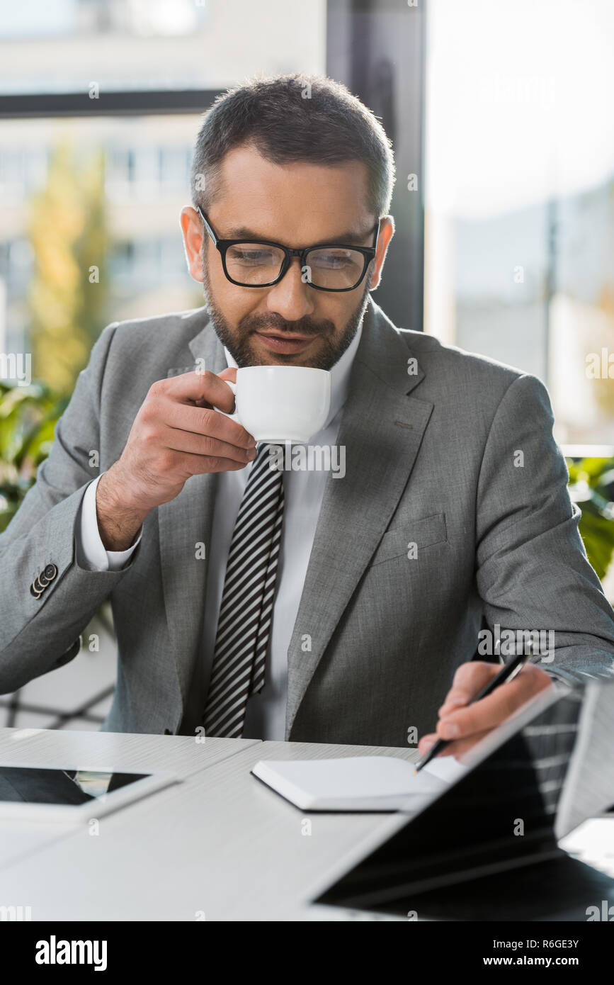 businessman in suit and eyeglasses drinking coffee and writing in notebook at wokrplace Stock Photo