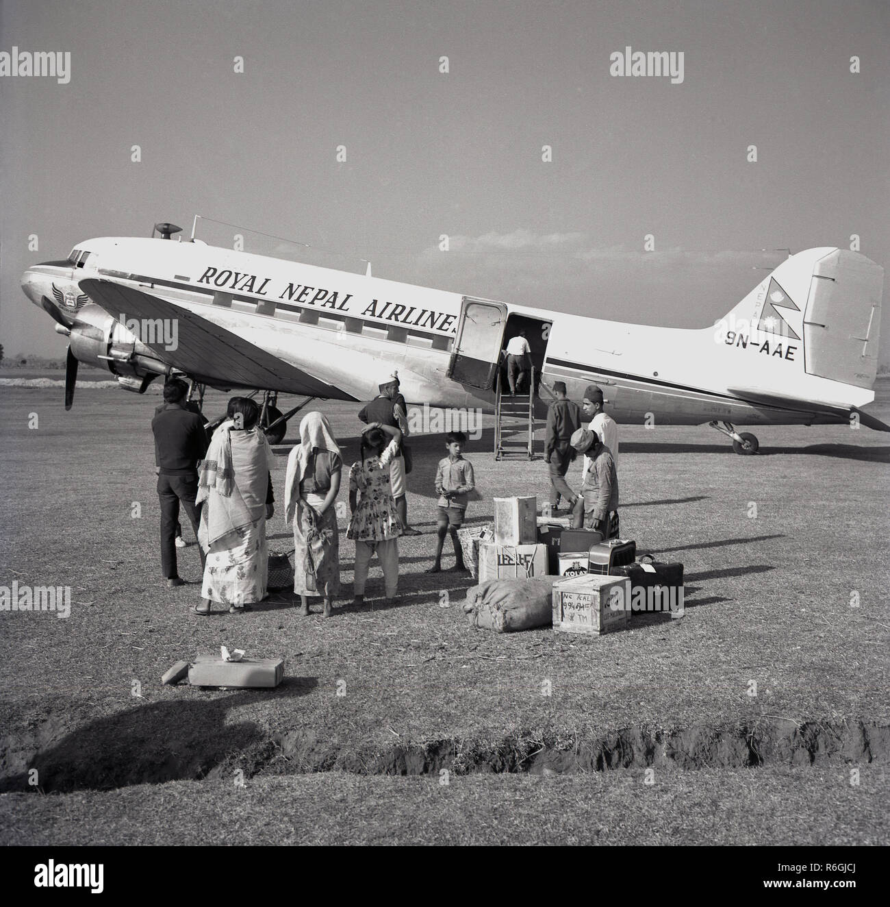 1959, historical, passengers standing outside with their luggage waiting to board a nearby propeller-driven Royal Nepal Airlines Douglas-DC3 aircraft parked on a grassy runway at Kathmandu, Nepal. Established in 1958, the DC-3 was the airline's first aircraft and used to serve domestic routes and destinations in India. Stock Photo