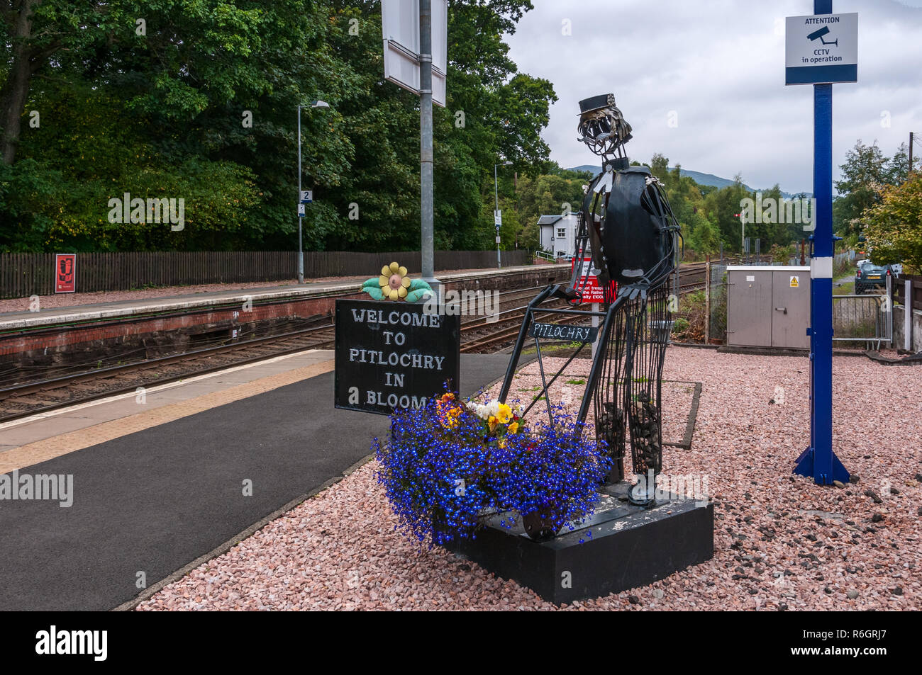 A quirky figure of a railway porter artfully sculpted from wire rods pushing a hand trolley bearing a variety of colourful blooming flowers Stock Photo