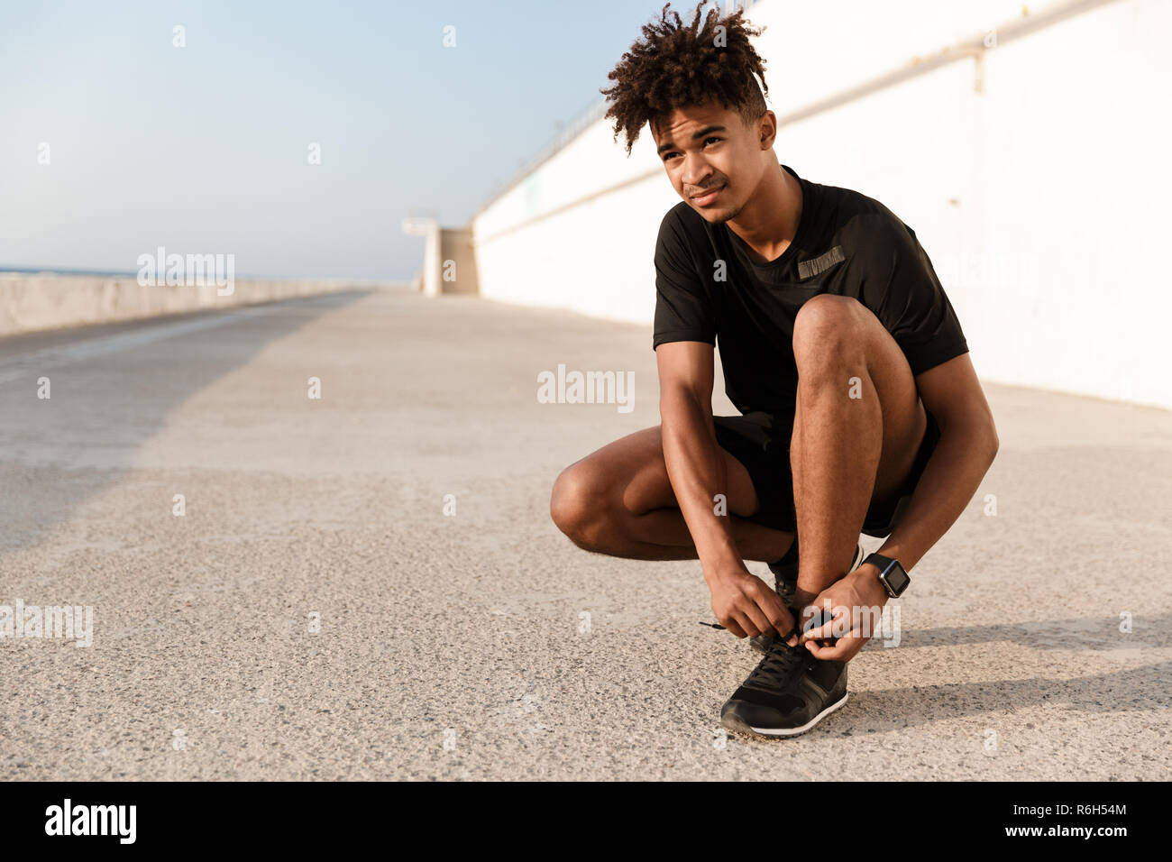 Strong young african man dressed in sportswear tying his shoelace at the beach Stock Photo