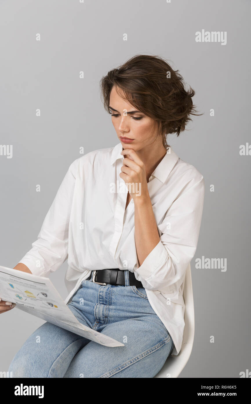 Photo Of A Thinking Young Woman Sitting On Stool Isolated Over Grey Background Holding Newspaper Reading Stock Photo Alamy