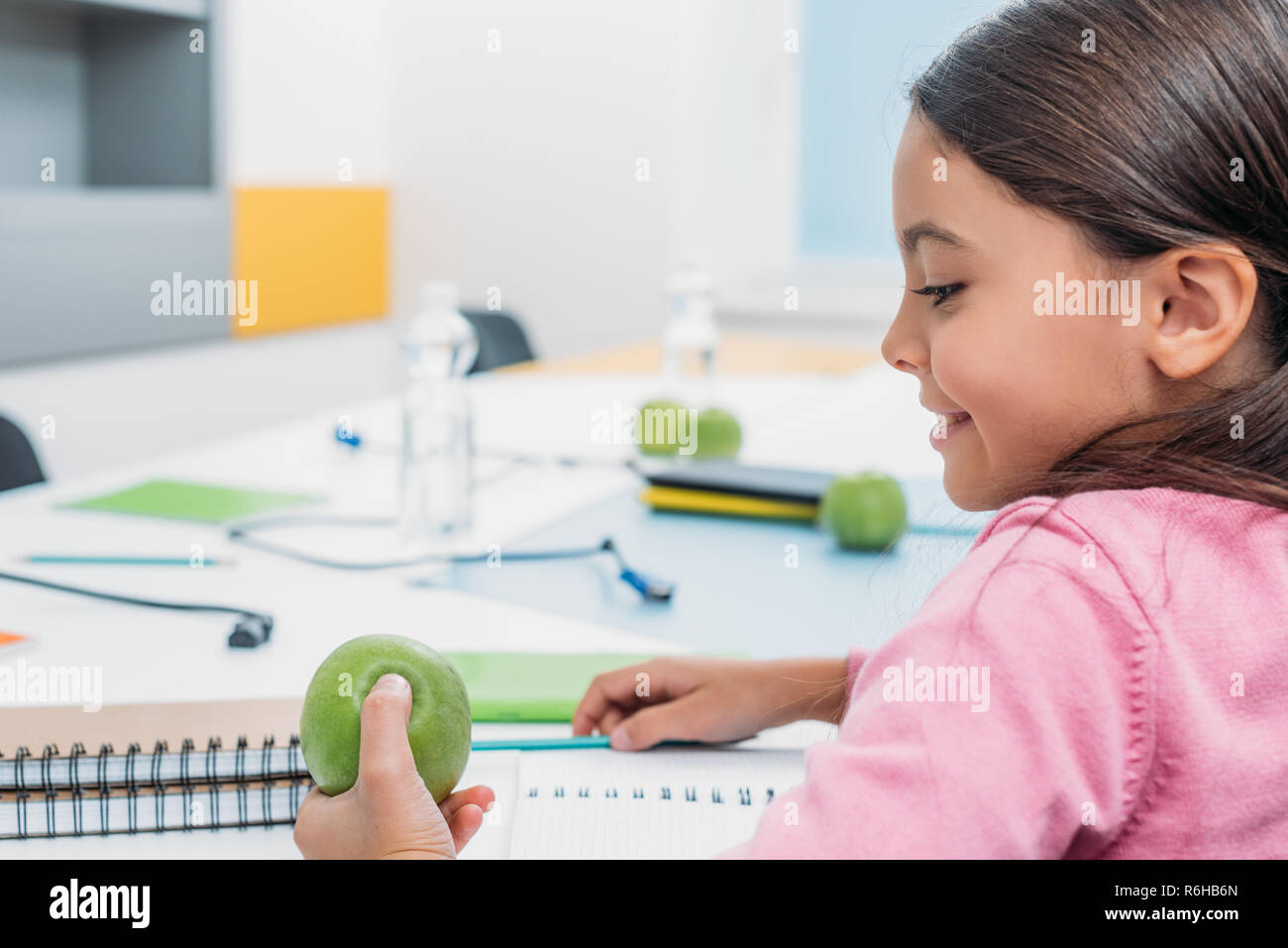 Adorable schoolgirl looking at apple while sitting at desk in classroom ...