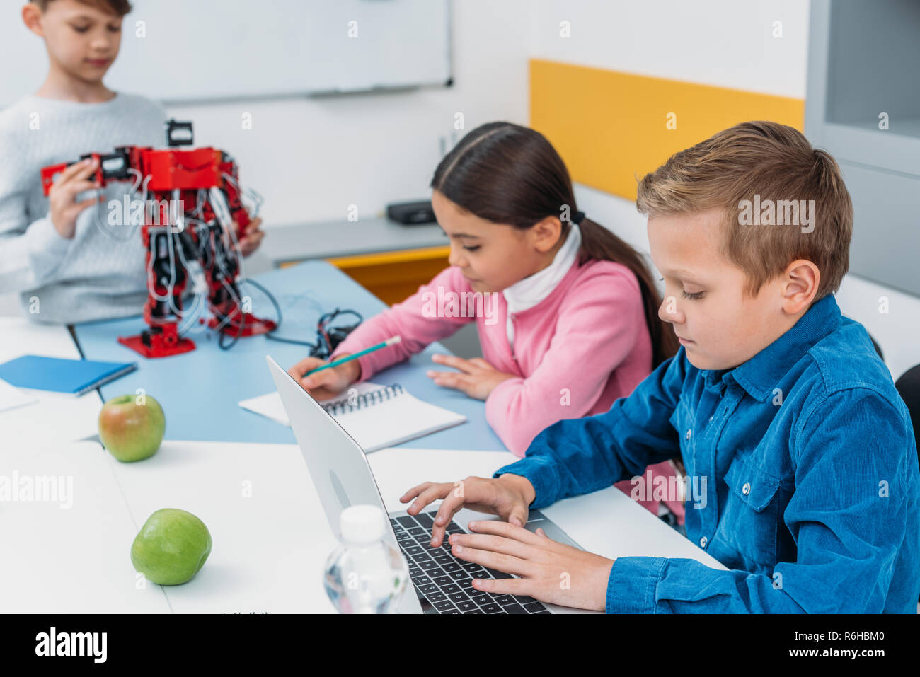 children writing in notebooks, typing laptop and touching red robot in ...