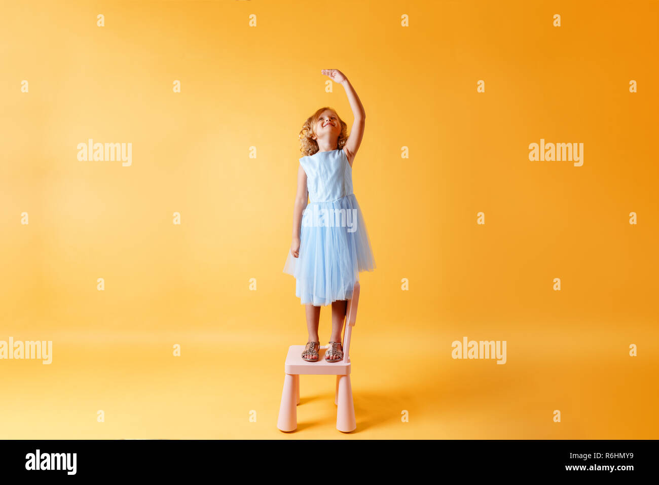 A little child girl in a blue dress is standing on a chair and measures her height against the background of the yellow wall. Concept of development,  Stock Photo
