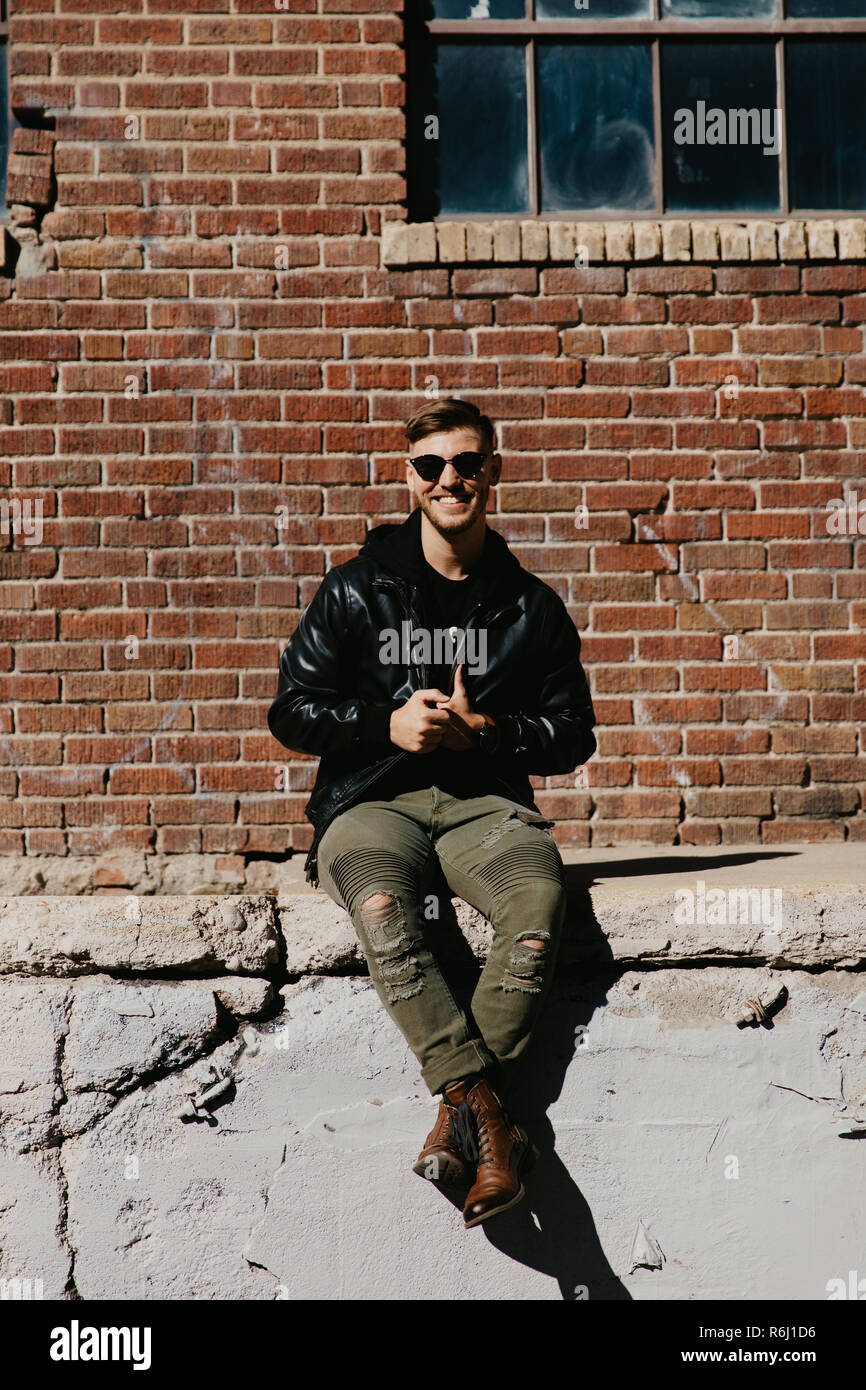 Young Attractive Modern Fashion Caucasian Male Guy Walking, Sitting, Smiling, and Laughing Outside Urban Old Abandoned Brick Urban Warehouse Building Stock Photo