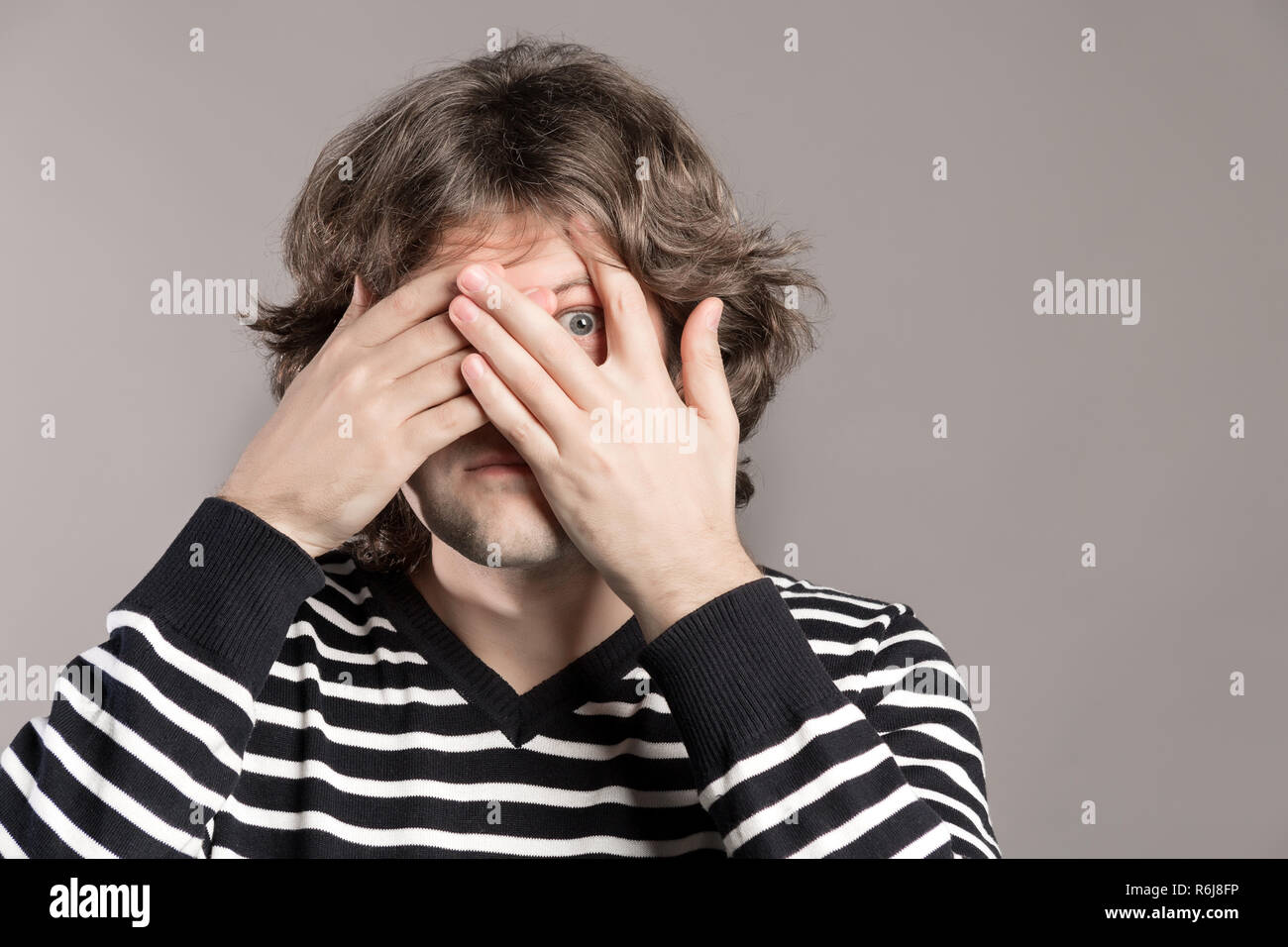 Young hipster man peeking through fingers. Shaggy male covers face with both hands, has frightened expression as notices something terrible or scarying over grey background. Embarrassed expression Stock Photo