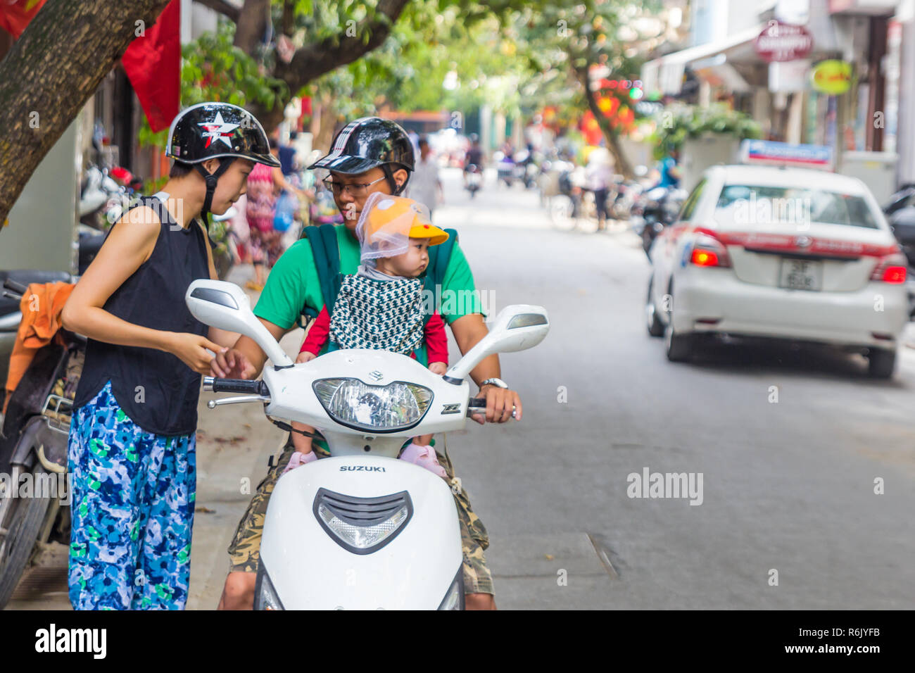 Hanoi, Vietnam - CIRCA October 2015: woman, man in helmets, child in baby carrier and yellow cap travel on scooter in Hanoi, Vietnam Stock Photo