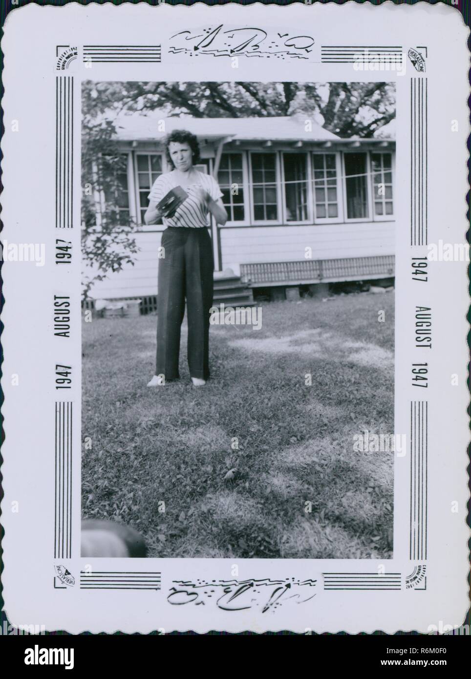 Woman standing on the lawn of a suburban home and holding a Kodak Brownie box camera, an iconic early 20th century camera, printed with an ornate decorated frame, August, 1947. () Stock Photo