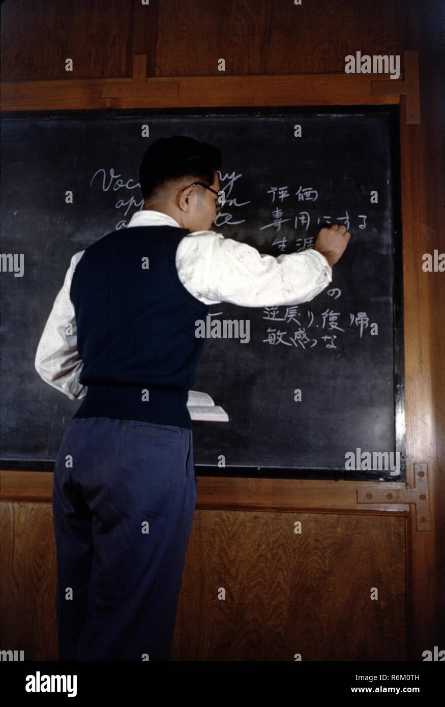 Color photograph of a Japanese man, seen from behind, wearing a dark vest and trousers and a white long-sleeved shirt, standing in front of a blackboard, holding an open book, and writing the Japanese equivalents for English vocabulary words on the board with a piece of chalk, likely photographed in Japan during the mid-twentieth century, 1965. () Stock Photo