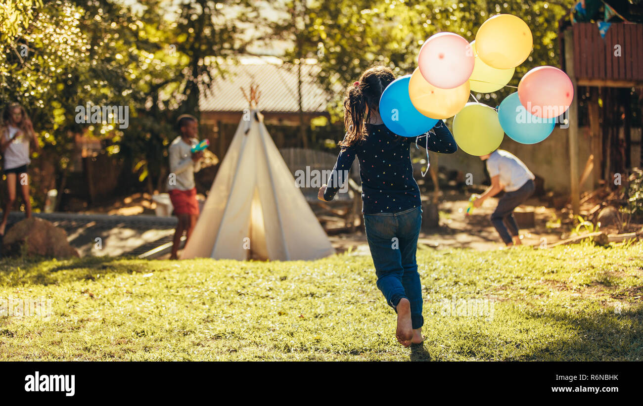Little girl with colorful balloons running towards kids playing in backyard. Children having fun outdoors. Stock Photo