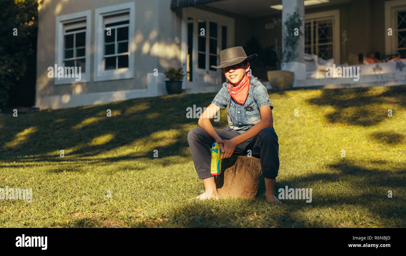 Little cowboy sitting outdoors in backyard with water pistol. Young boy wearing cowboy hat with water squirt gun. Stock Photo