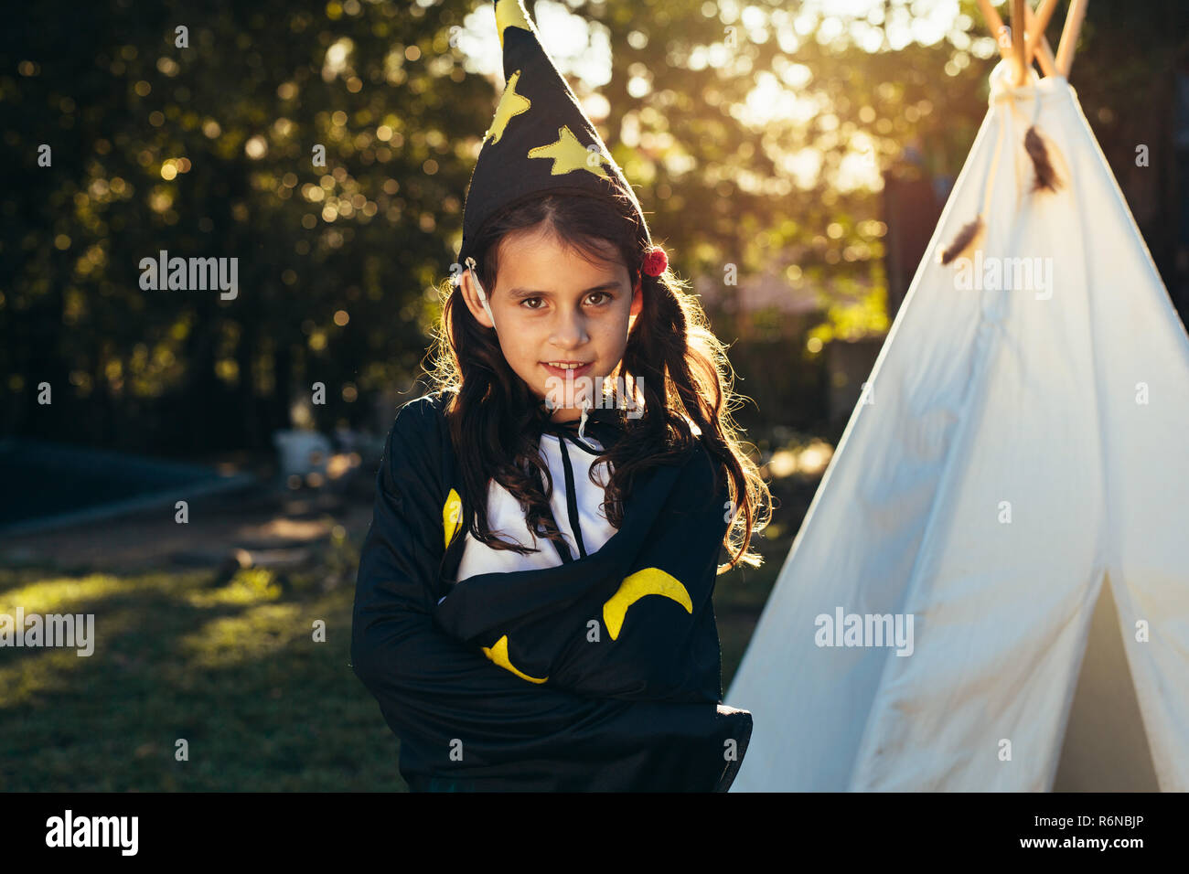 Cute little girl in wizard costume wearing hat with her arms crossed. Cute young little girl in halloween dress standing in backyard garden teepee in Stock Photo