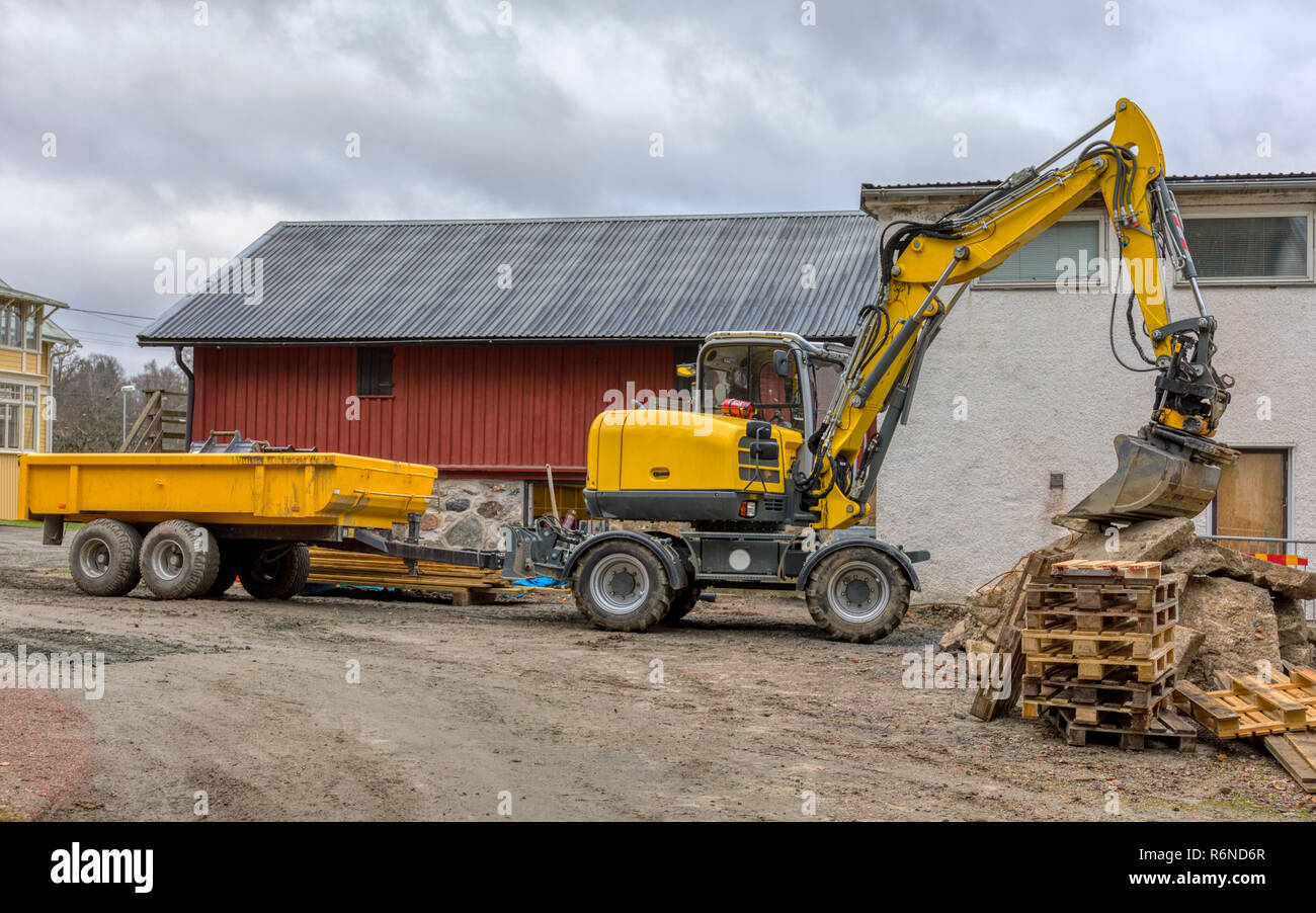FLODA, SWEDEN - NOVEMBER 21 2018: Medium sized yellow excavator with trailer resting it's bucket on pile of wood pallets Stock Photo