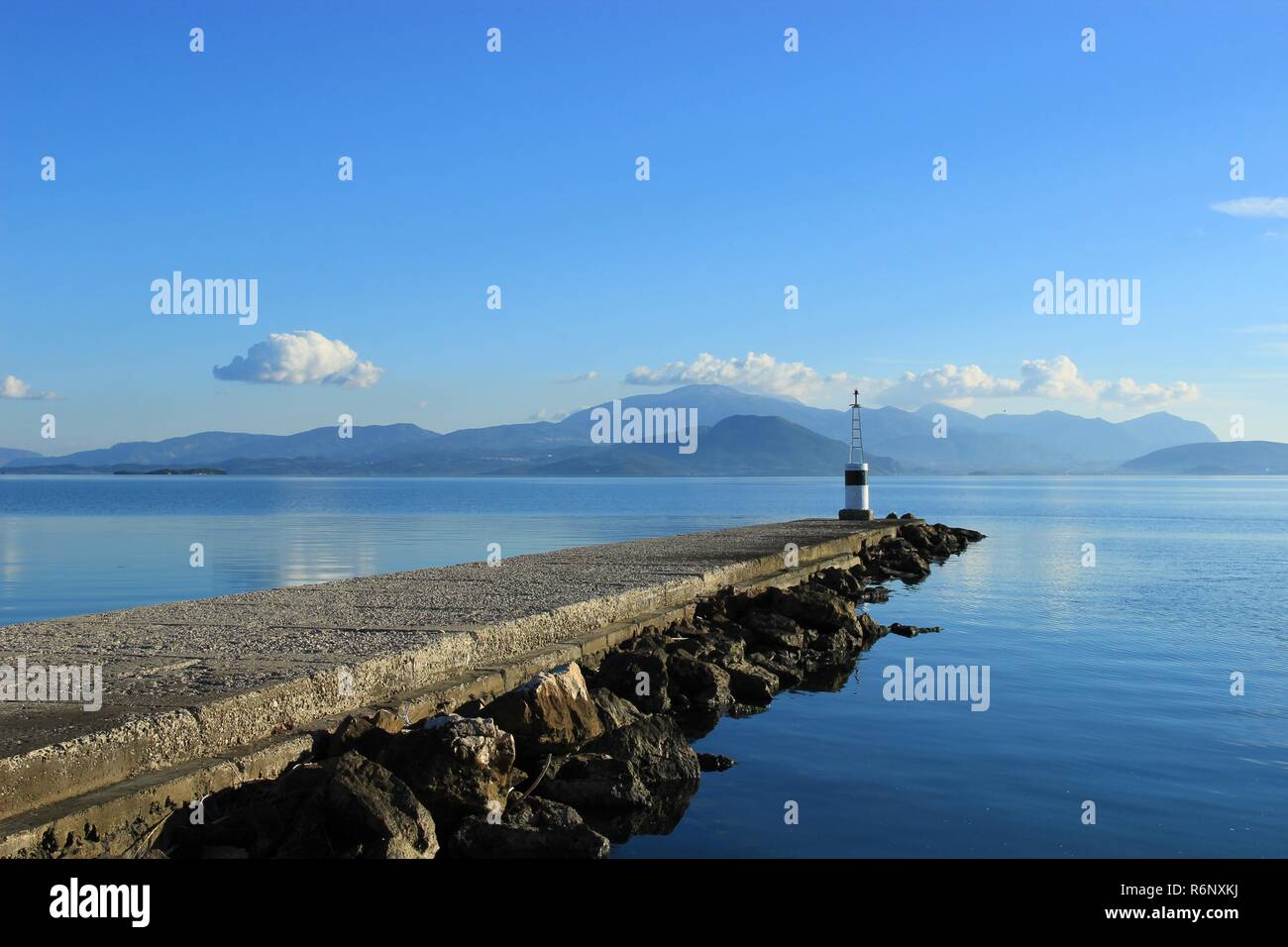 Lighthouse at the end of pier at Koronisia village in Ambracian gulf, Epirus Greece, calm blue sea, clouds in the sky and view of mountains and hills Stock Photo