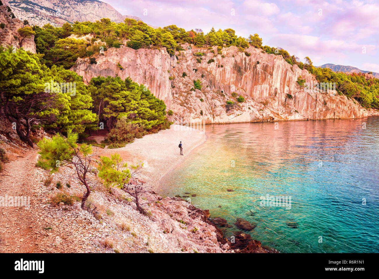 young man with backpack standing on empty secluded Nugal beach in Makarska Riviera, Croatia after sunset looking at sea Stock Photo